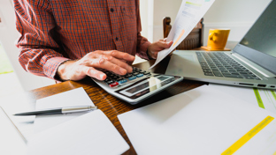 A person in a red plaid shirt is seated at a wooden table, working on financial tasks. They are using a large calculator with one hand while holding a printed bill or financial statement in the other.