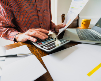 A person in a red plaid shirt is seated at a wooden table, working on financial tasks. They are using a large calculator with one hand while holding a printed bill or financial statement in the other.