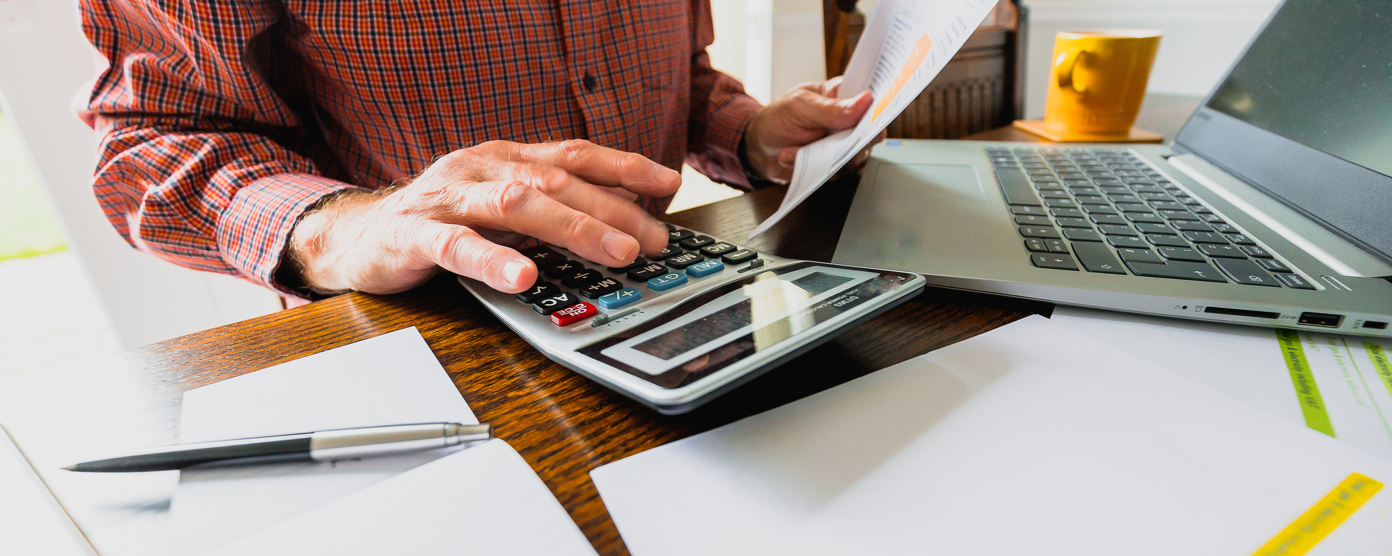 A person in a red plaid shirt is seated at a wooden table, working on financial tasks. They are using a large calculator with one hand while holding a printed bill or financial statement in the other.