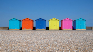 A row of brightly painted beach huts stands on sandy ground, each in different pastel and vibrant colors.