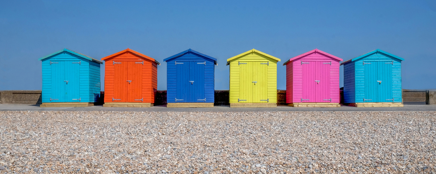 A row of brightly painted beach huts stands on sandy ground, each in different pastel and vibrant colors.