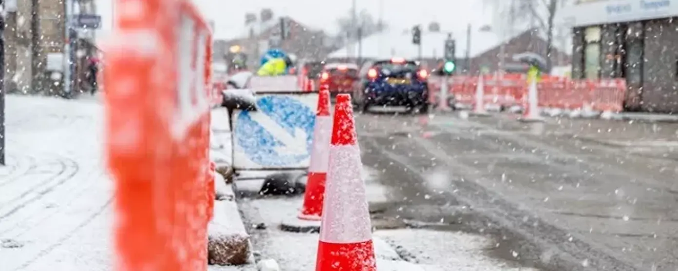 A snowy street scene shows bright orange traffic cones and barriers lining a construction area along the roadside.