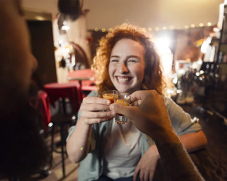 A smiling young woman with curly hair toasting a friend with a shot in a pub
