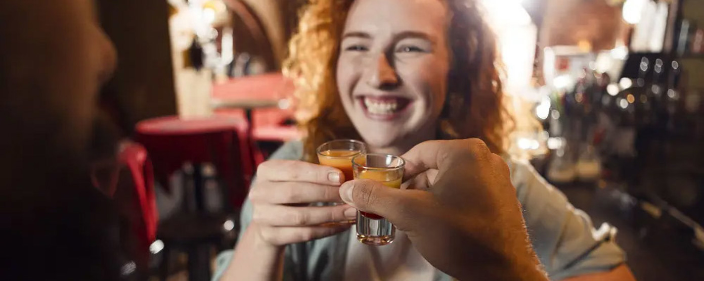 A smiling young woman with curly hair toasting a friend with a shot in a pub