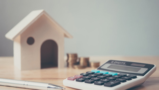 A calculator, pen, coin stacks, and a house model represent home finance, budgeting, and property investment calculations.