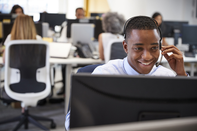 A young man wearing a headset is smiling and speaking while working at a computer in a busy office environment.