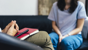 Close-up view of a therapy or counseling session, showing a therapist with a notebook and a client sitting across