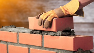 A close-up image of a construction worker wearing protective orange gloves carefully placing a red brick onto wet mortar atop a partially built brick wall.