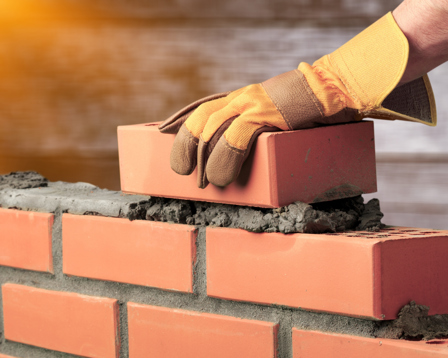 A close-up image of a construction worker wearing protective orange gloves carefully placing a red brick onto wet mortar atop a partially built brick wall.