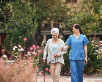 An elderly woman using a cane walks through a garden with pink flowers, supported by a smiling caregiver in blue scrubs