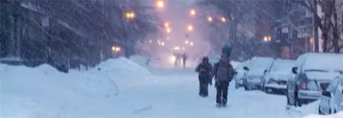 A snow-covered city street is seen during a heavy snowstorm, with visibility reduced due to falling snow.