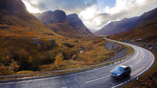 A vehicle travels along a curvy mountain road, surrounded by hills showcasing rich autumn foliage.
