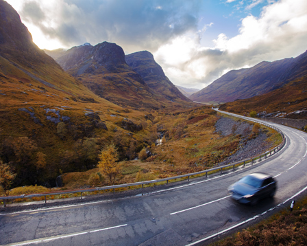 A vehicle travels along a curvy mountain road, surrounded by hills showcasing rich autumn foliage.