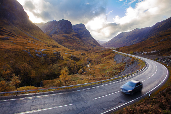 A vehicle travels along a curvy mountain road, surrounded by hills showcasing rich autumn foliage.