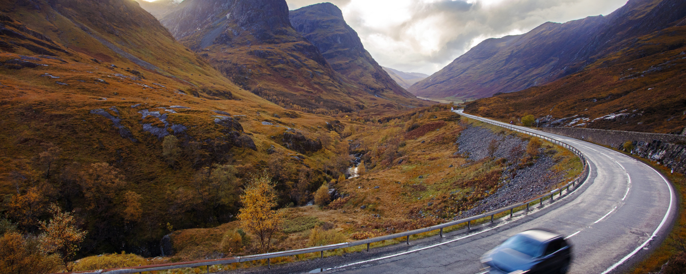 A vehicle travels along a curvy mountain road, surrounded by hills showcasing rich autumn foliage.
