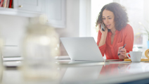 A woman in a red shirt works from home, speaking on the phone while using a laptop, with a coffee cup and open binder on the table