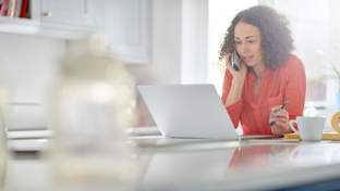 A woman in a red shirt works from home, speaking on the phone while using a laptop, with a coffee cup and open binder on the table