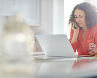 A woman in a red shirt works from home, speaking on the phone while using a laptop, with a coffee cup and open binder on the table