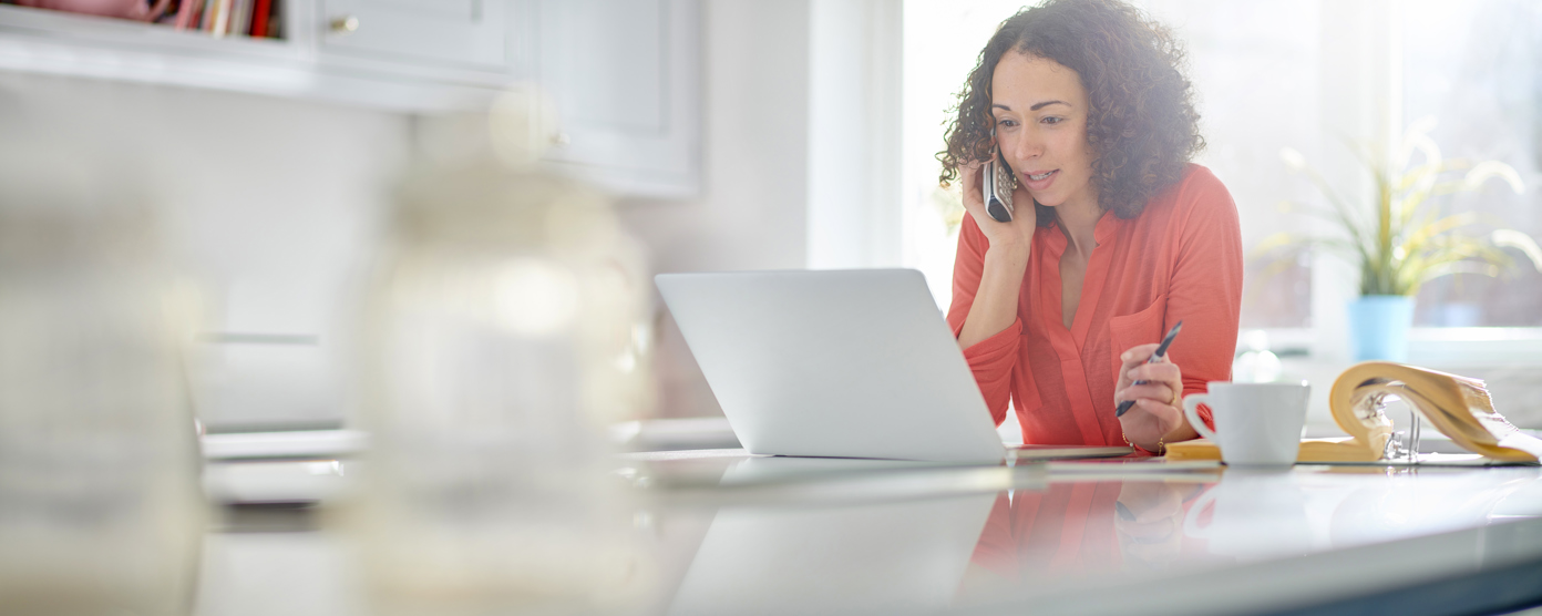 A woman in a red shirt works from home, speaking on the phone while using a laptop, with a coffee cup and open binder on the table