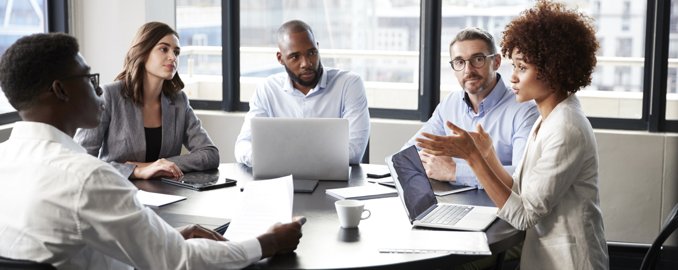 A group of five diverse professionals engaged in a business meeting around a round table in a modern office.