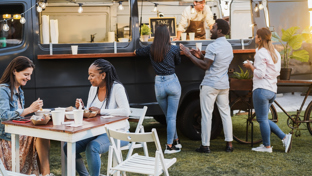 People order from a dark food truck while friends eat at nearby outdoor tables on a grassy area