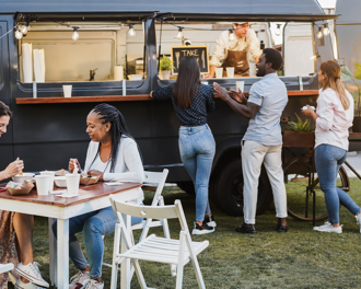 People order from a dark food truck while friends eat at nearby outdoor tables on a grassy area