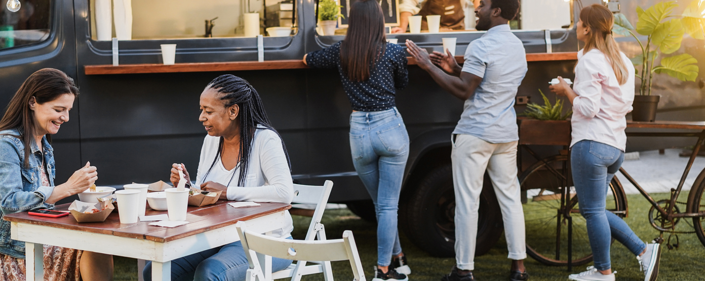 People order from a dark food truck while friends eat at nearby outdoor tables on a grassy area