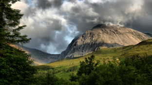 Dramatic view of a rocky mountain under stormy clouds, overlooking a green valley and forest.