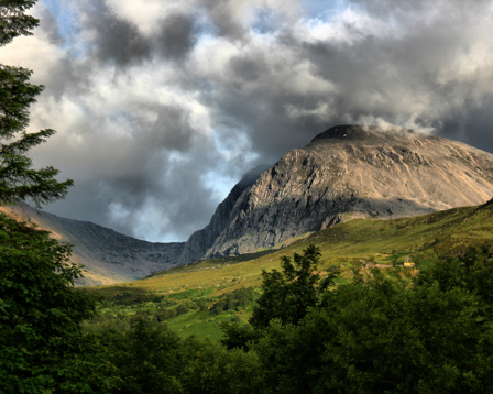 Dramatic view of a rocky mountain under stormy clouds, overlooking a green valley and forest.