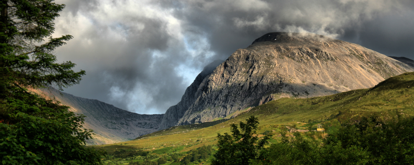Dramatic view of a rocky mountain under stormy clouds, overlooking a green valley and forest.