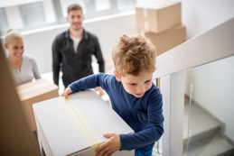 A young boy carries a moving box up a staircase, followed by a smiling adult woman and a man, indicating a family moving into a new home.