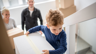 A young boy carries a moving box up a staircase, followed by a smiling adult woman and a man, indicating a family moving into a new home.