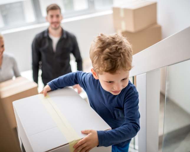 A young boy carries a moving box up a staircase, followed by a smiling adult woman and a man, indicating a family moving into a new home.
