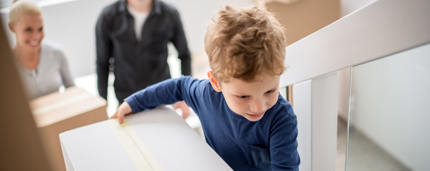 A young boy carries a moving box up a staircase, followed by a smiling adult woman and a man, indicating a family moving into a new home.