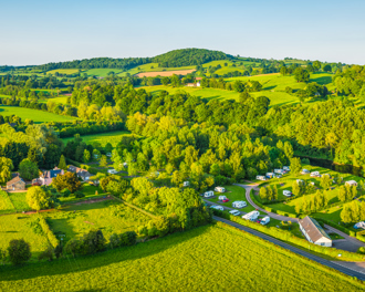 A panoramic aerial view of a vibrant green countryside landscape, featuring gently rolling hills, clusters of trees, and wide open fields.