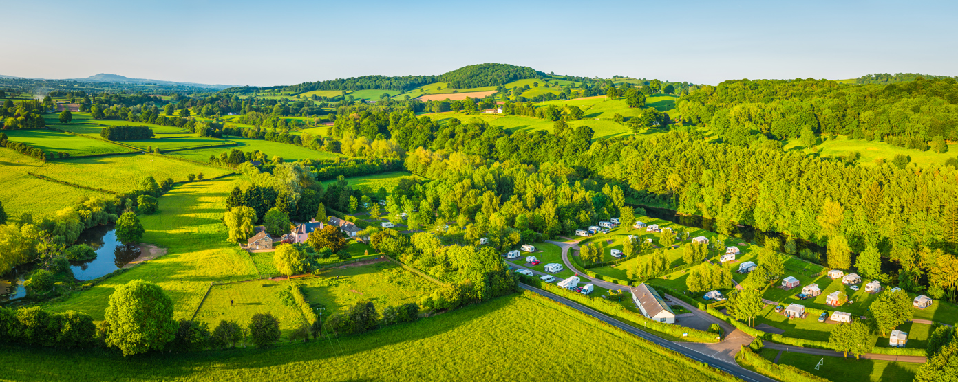 A panoramic aerial view of a vibrant green countryside landscape, featuring gently rolling hills, clusters of trees, and wide open fields.