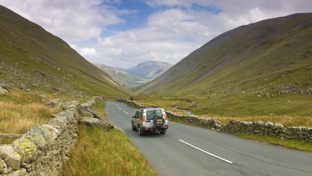 SUV driving along a narrow mountain pass in the Lake District with dry-stone walls and sweeping valley views