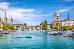 Scenic Swiss city riverfront with churches, bridges, boats, and a bright blue sky.