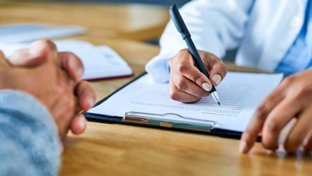 Close-up of a clinician writing on a clipboard during a consultation, with a patient’s clasped hands visible across the desk.