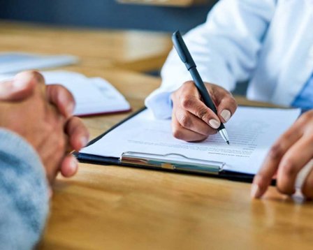 Close-up of a clinician writing on a clipboard during a consultation, with a patient’s clasped hands visible across the desk.