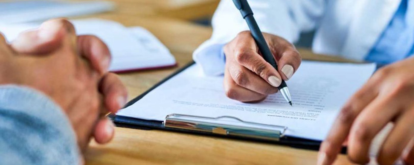 Close-up of a clinician writing on a clipboard during a consultation, with a patient’s clasped hands visible across the desk.