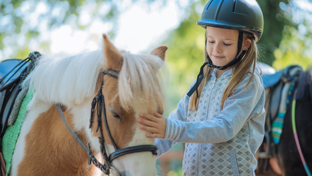 A girl in a riding helmet stands beside a saddled pony, typical of equestrian settings for teaching young riders.