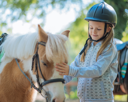 A girl in a riding helmet stands beside a saddled pony, typical of equestrian settings for teaching young riders.
