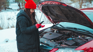 In a snowy setting, a woman in winter attire examines her phone next to her car with the hood raised and engine trouble.