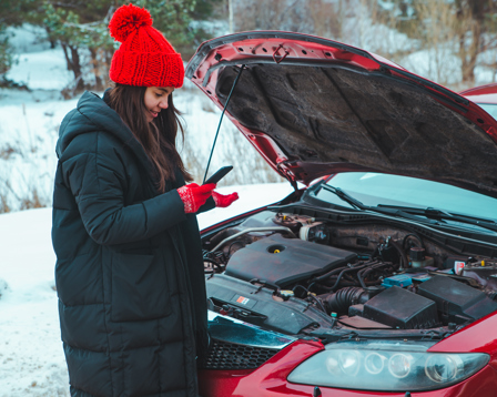 In a snowy setting, a woman in winter attire examines her phone next to her car with the hood raised and engine trouble.