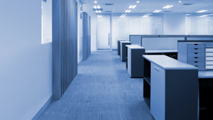 A clean and modern open-plan office space with a blue color tone. The area features carpeted flooring, bright overhead lighting, and rows of empty workstations with drawers and cabinets.