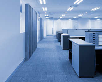 A clean and modern open-plan office space with a blue color tone. The area features carpeted flooring, bright overhead lighting, and rows of empty workstations with drawers and cabinets.
