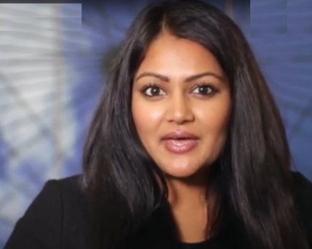 A woman with long, dark hair and medium skin tone is speaking or presenting indoors.