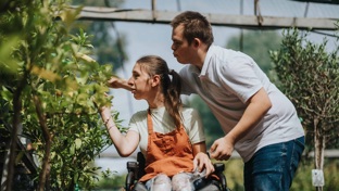 A male carer looks at a flower bush with a lady in a wheelchair