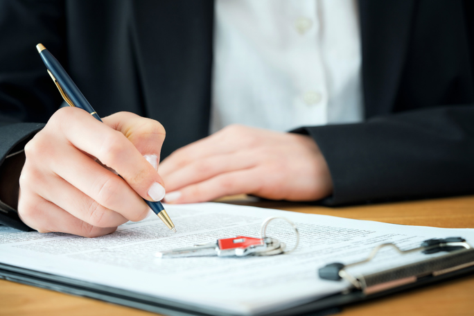 Close-up of hands signing a document with house keys, suggesting a formal real estate agreement.